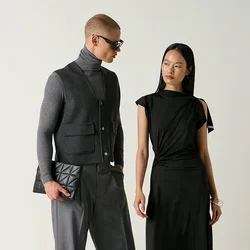 Homme en pull à col roulé gris, gilet et pantalon tenant une pochette, et femme en robe noire et chaussures debout près de chaises, dont une est renversée sur le sol.