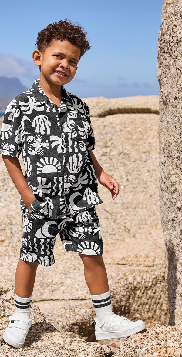 Junge trägt kurzärmliges Hemd, kurze Hose, schwarz weiß gemustert, weiße Sneakers, gestreifte Socken, Vorderansicht