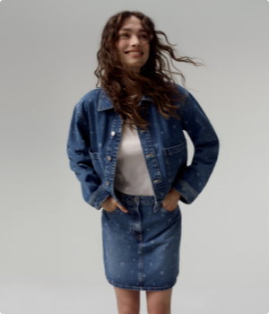 Frau trägt TOM TAILOR Denim Jeansjacke ohne Kapuze mit All-Over-Herz-Print in mid blue und passenden TOM TAILOR Denim Jeansrock mit All-Over Print in mid blue laser hearts denim