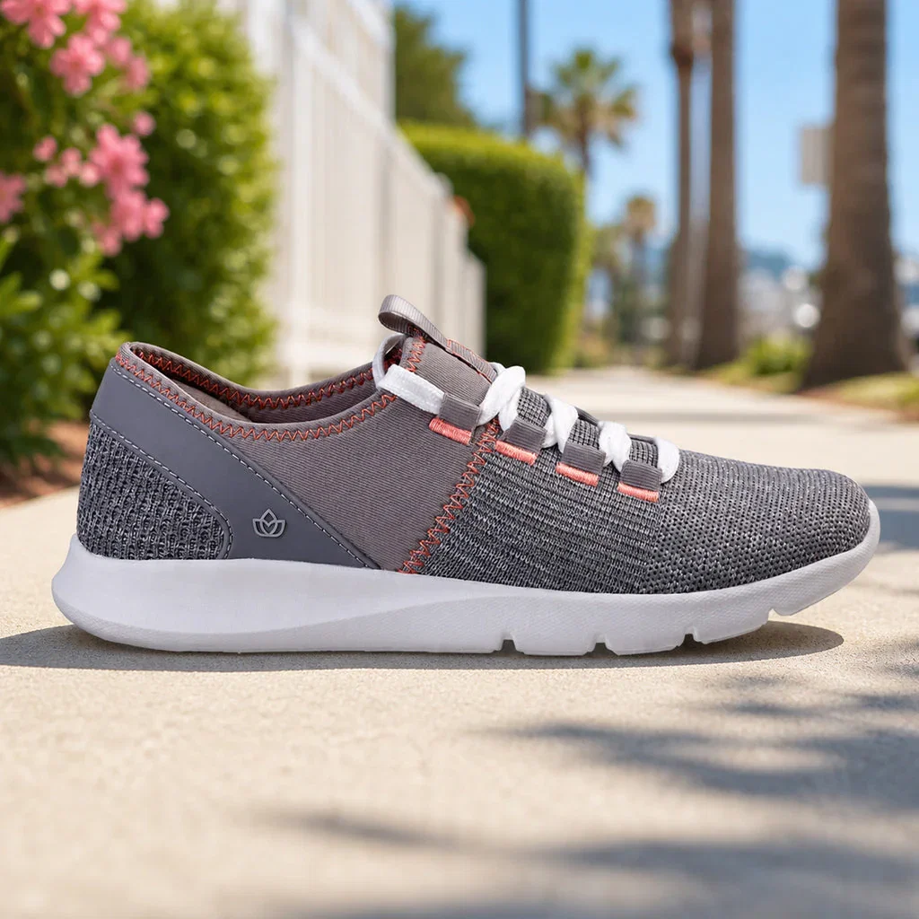 Gray knit sneaker with coral accents and white sole on sunny street with palm trees and pink flowers.