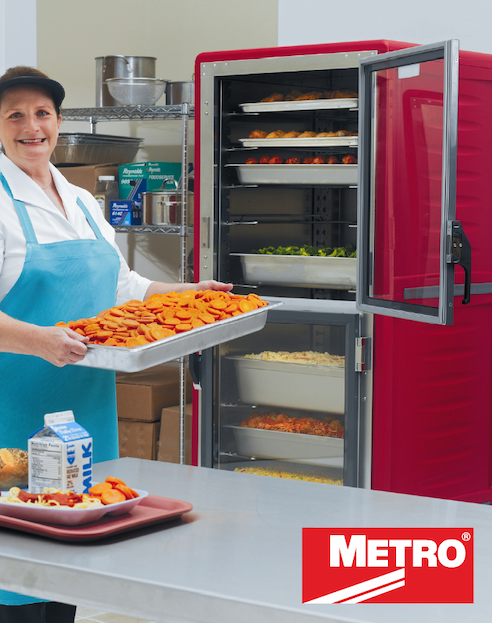 Metro heated holding cabinet with open door showing multiple trays of food, as a staff member places a tray of sliced vegetables inside.