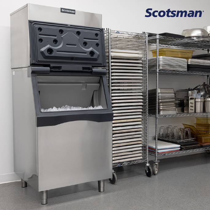 Scotsman commercial ice machine with open storage bin filled with ice, placed beside a metal rack with stacked trays in a kitchen prep area.