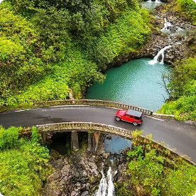 An aerial view of a red jeep driving over a bridge through a lush, tropical forest with waterfalls and a turquoise pool.