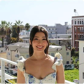 A portrait of a smiling woman on a balcony with a city skyline and palm trees in the background.