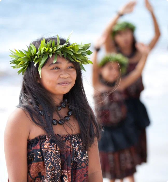 A young girl wearing a traditional haku lei and Hawaiian attire participating in a hula dance on the beach.