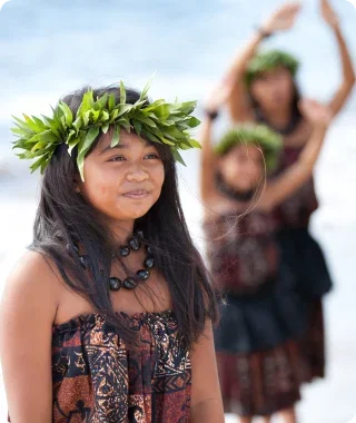 A young girl wearing a traditional haku lei and Hawaiian attire participating in a hula dance on the beach.
