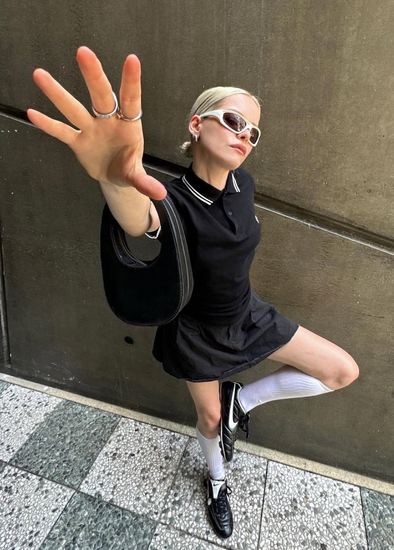 Taken from Instagram: a woman with blonde hair puts her hand up towards the camera and wears the Fred Perry Shirt in Black / Champagne / Champagne with a black mini skirt and white rimmed sunglasses on