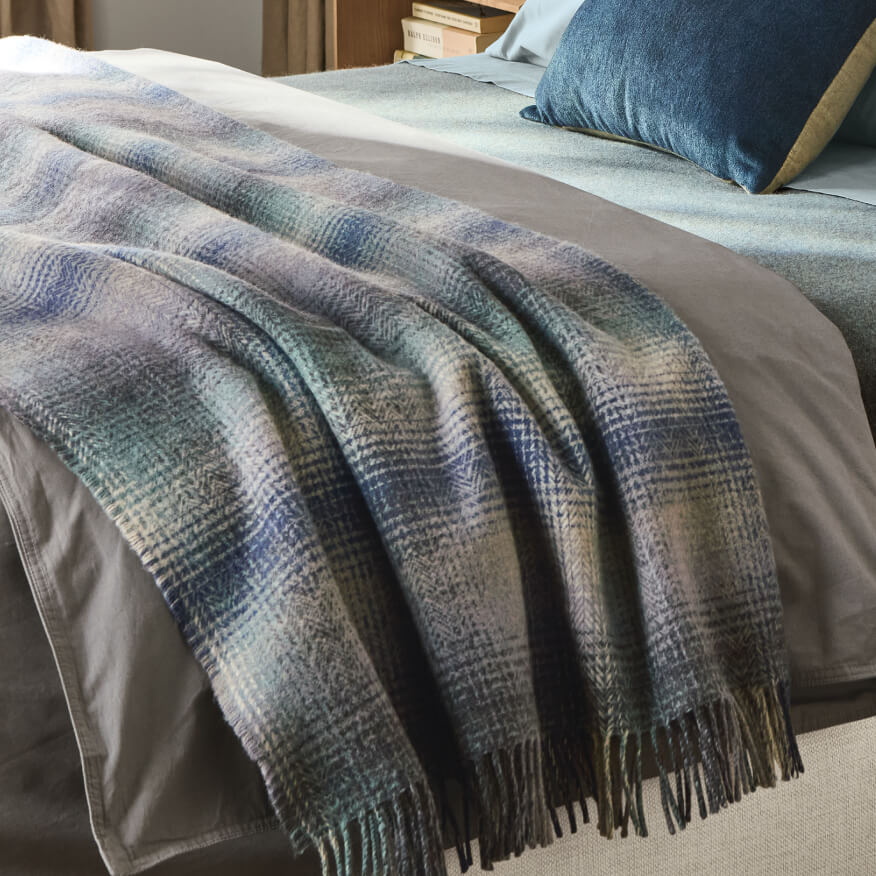 Sutter throw with blue-grey plaid pattern, draped across a bed with pillows in the background.