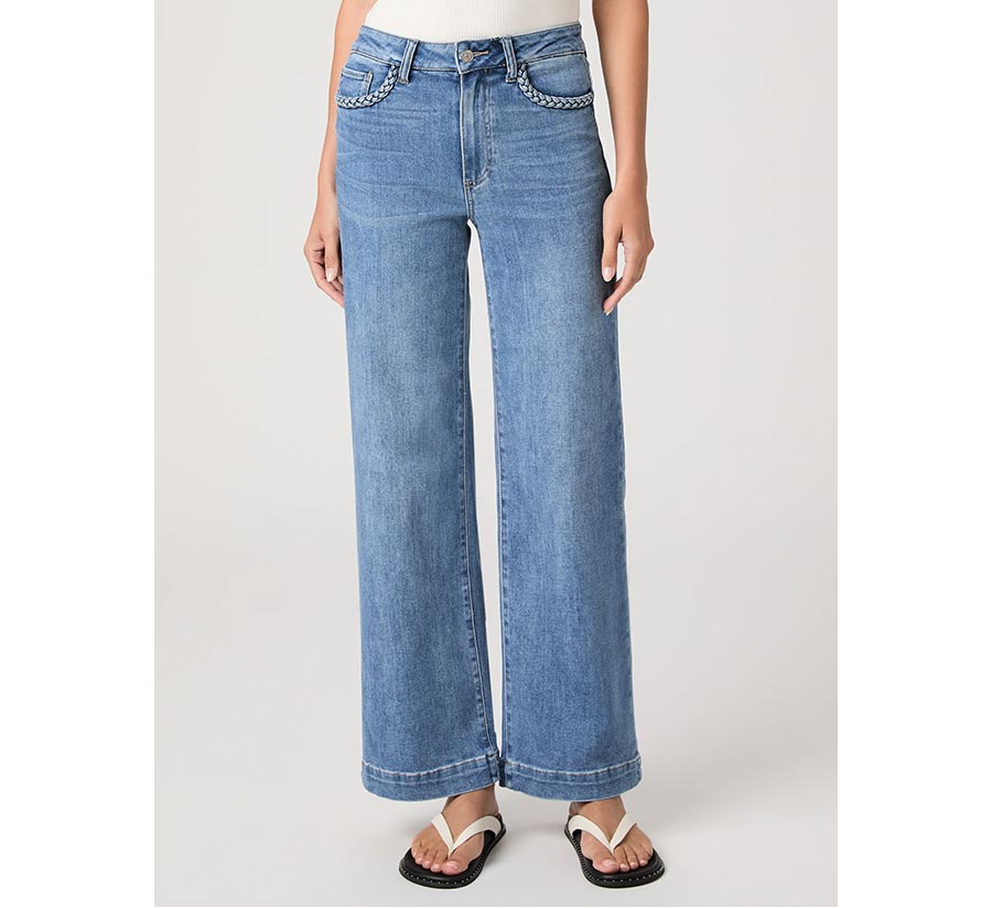 Woman wearing a pair of medium wash high-rise wide-leg jeans with braided detailing, a white tank, and leather cream and black flip-flop sandals, against a grey backdrop.
