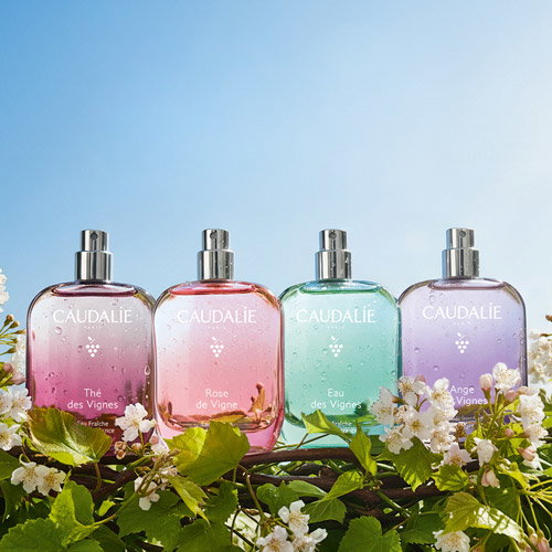 Four Caudalie bottles (pink, coral, mint, lavender) labelled Thé, Rose, Eau and Ange des Vignes among flowering vines, blue sky