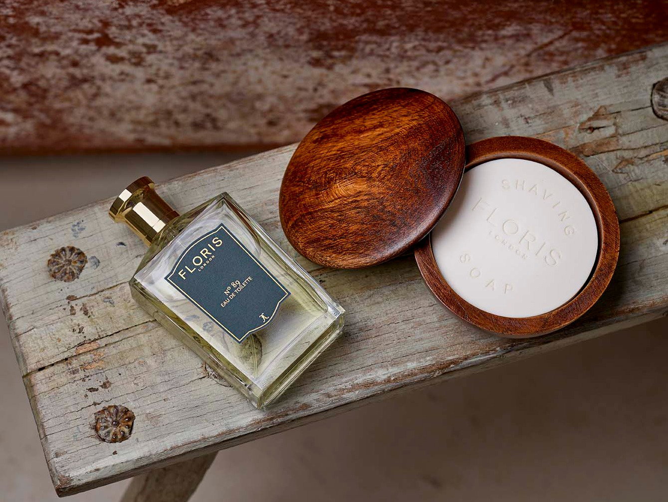 A bottle of Floris London Eau de Toilette and an open wooden shaving soap dish with soap sit on a weathered wooden bench.