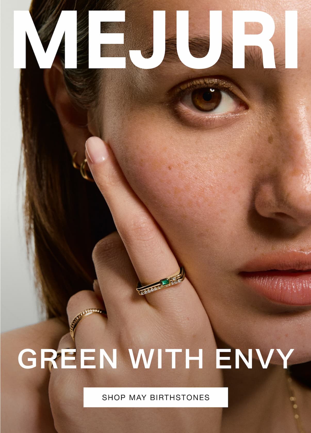 Woman wearing gold emerald rings and hoop earrings. Text reads: MEJURI. GREEN WITH ENVY. Shop May Birthstones.