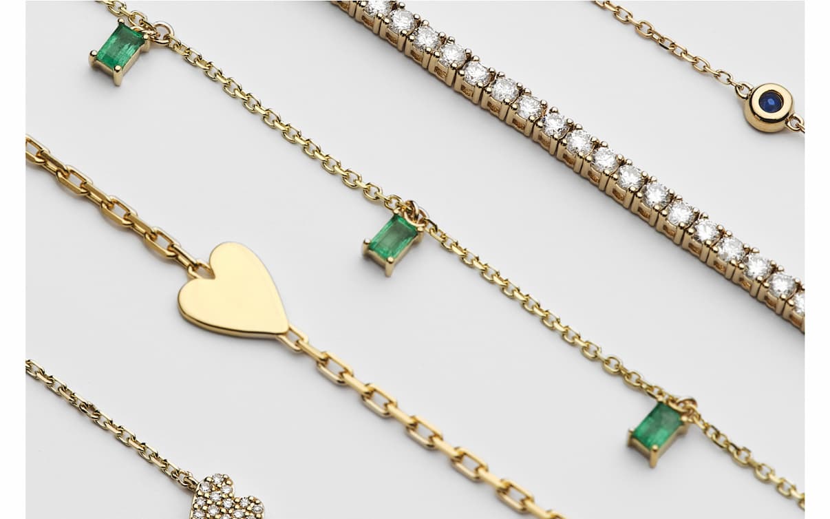 Gold chain bracelets with emerald, diamond, and heart charm details, styled on a light background.