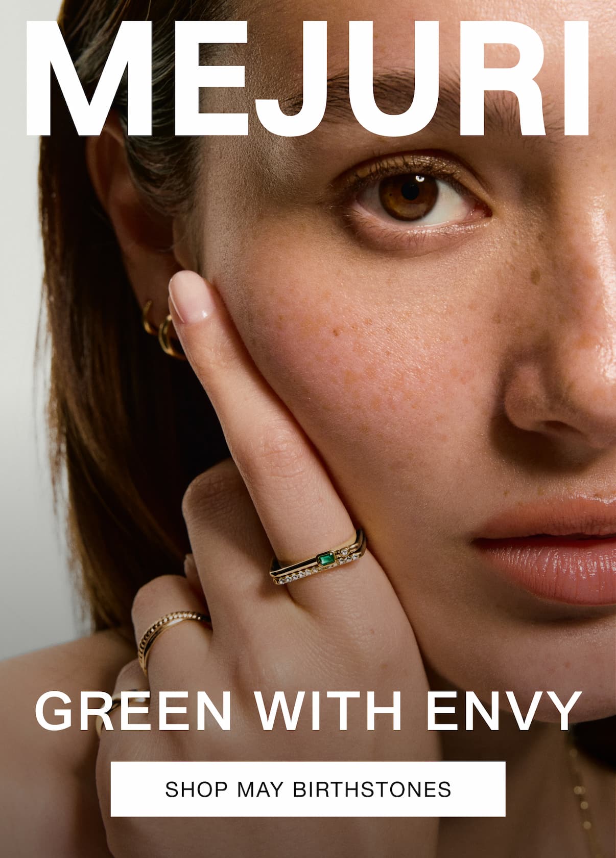 Woman wearing gold emerald rings and hoop earrings. Text reads: MEJURI. GREEN WITH ENVY. Shop May Birthstones.