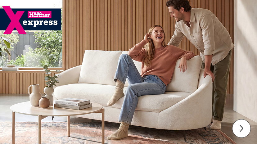 Ein Paar lacht gemeinsam in einem modern eingerichteten Wohnzimmer; die Frau sitzt auf einem cremeweißen Sofa, der Mann lehnt daneben. Oben links ist das Logo ‚Höffner Express‘ platziert.