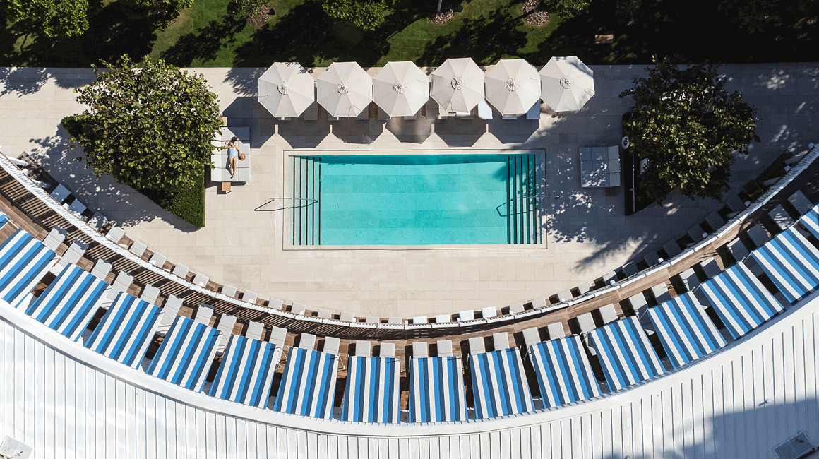 Top-down view of a rectangular pool lined with white umbrellas and blue-striped loungers in a curved terrace.