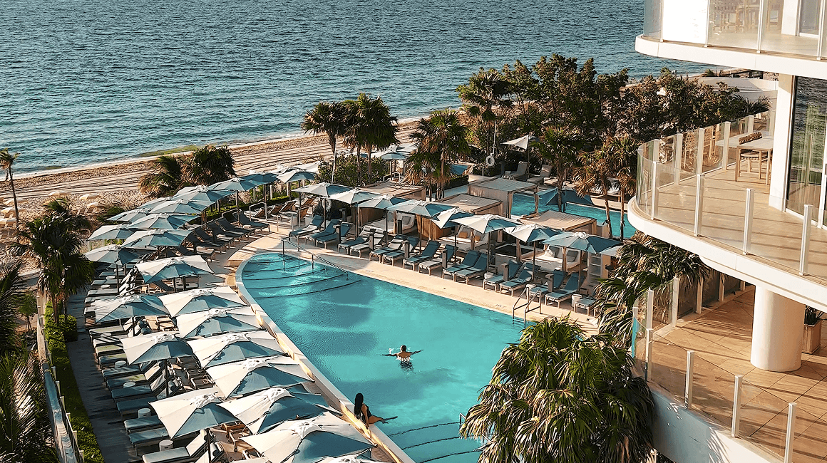 Oceanfront pool terrace with curved edges, loungers, umbrellas, and the shoreline beyond.