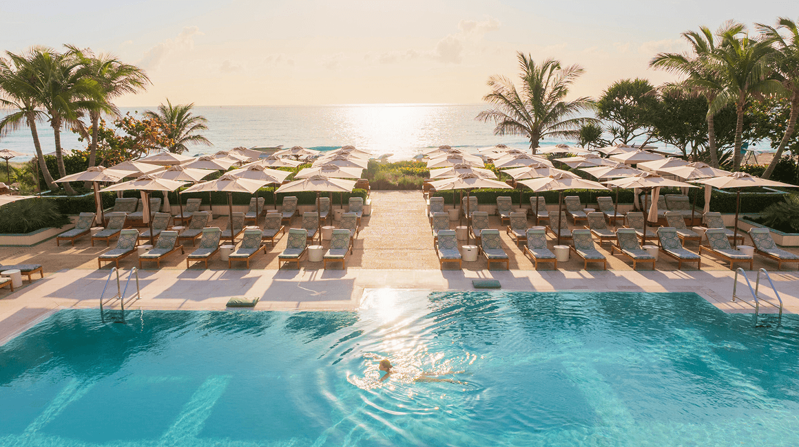 A woman swims in an infinity pool facing the ocean, with rows of loungers and palm trees.