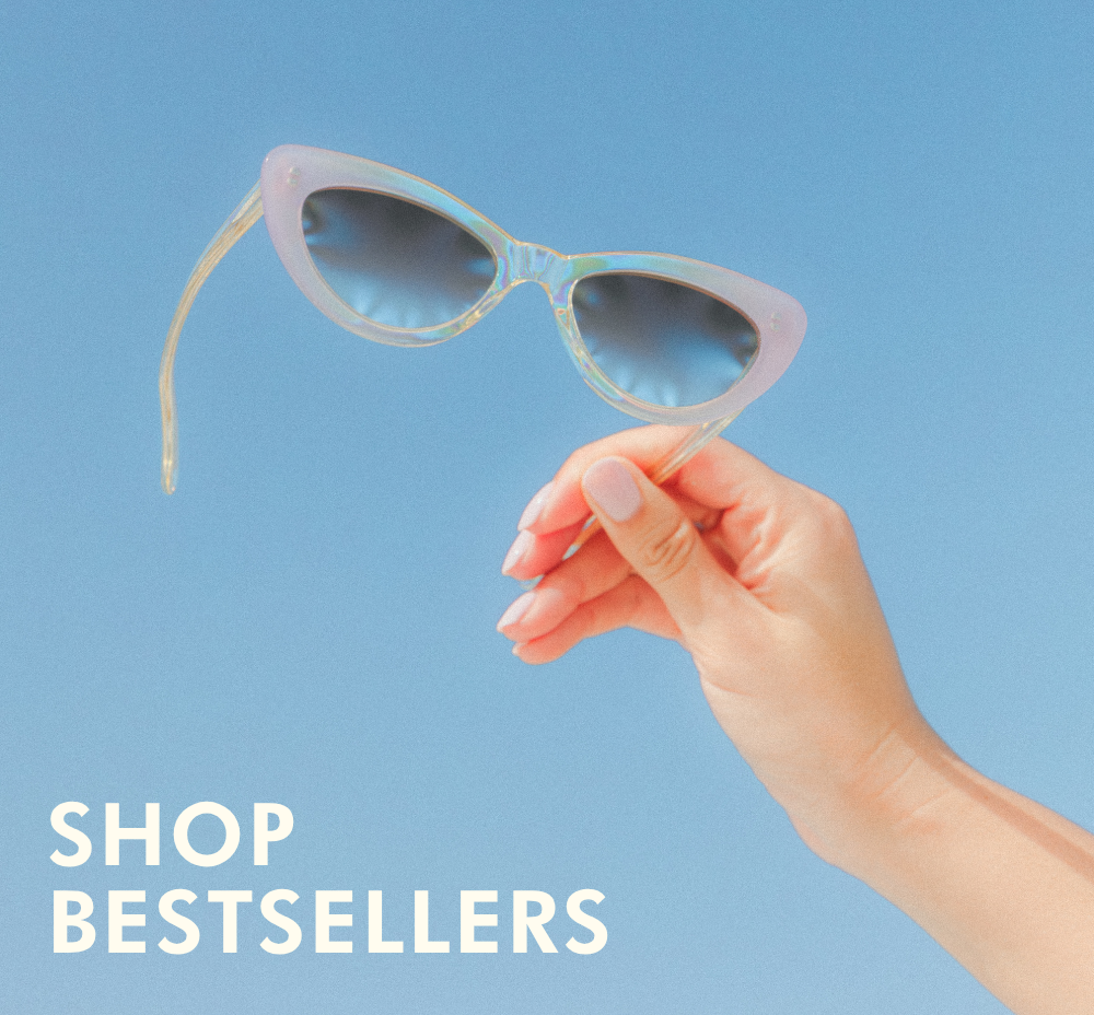 A hand with light pink manicured nails holds up a pair of pointed cat-eye sunglasses against a bright blue background. The frames are a translucent, iridescent light pink that catches the light, paired with blue gradient lenses. Bold white text in the bottom left corner reads: 