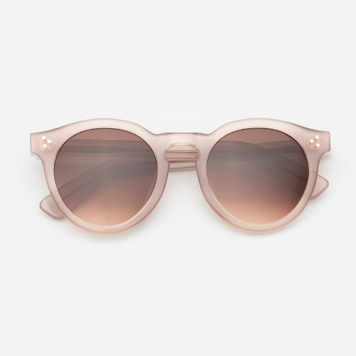 A studio product shot of round sunglasses in a frosted, translucent pale pink acetate. The lenses are a warm brown gradient, and the temples feature a three-dot silver pin detail.