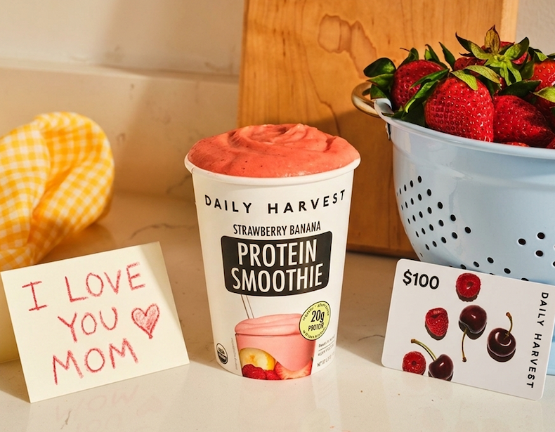 Daily Harvest Strawberry Banana protein smoothie on a kitchen counter with fresh strawberries, a $100 gift card, and an “I love you mom” note.