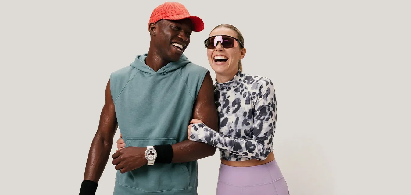 Hombre con sudadera sin mangas y gorra roja sonríe con mujer con top estampado y gafas de sol mientras caminan abrazados sobre un fondo liso.