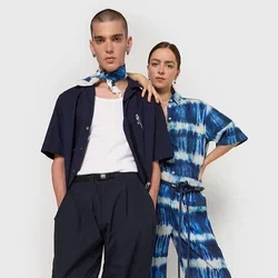 Twee modellen staan samen. De één draagt een marineblauw shirt en wijde broek; de ander heeft een blauw tie-dye jumpsuit aan. Beide hebben minimale accessoires.