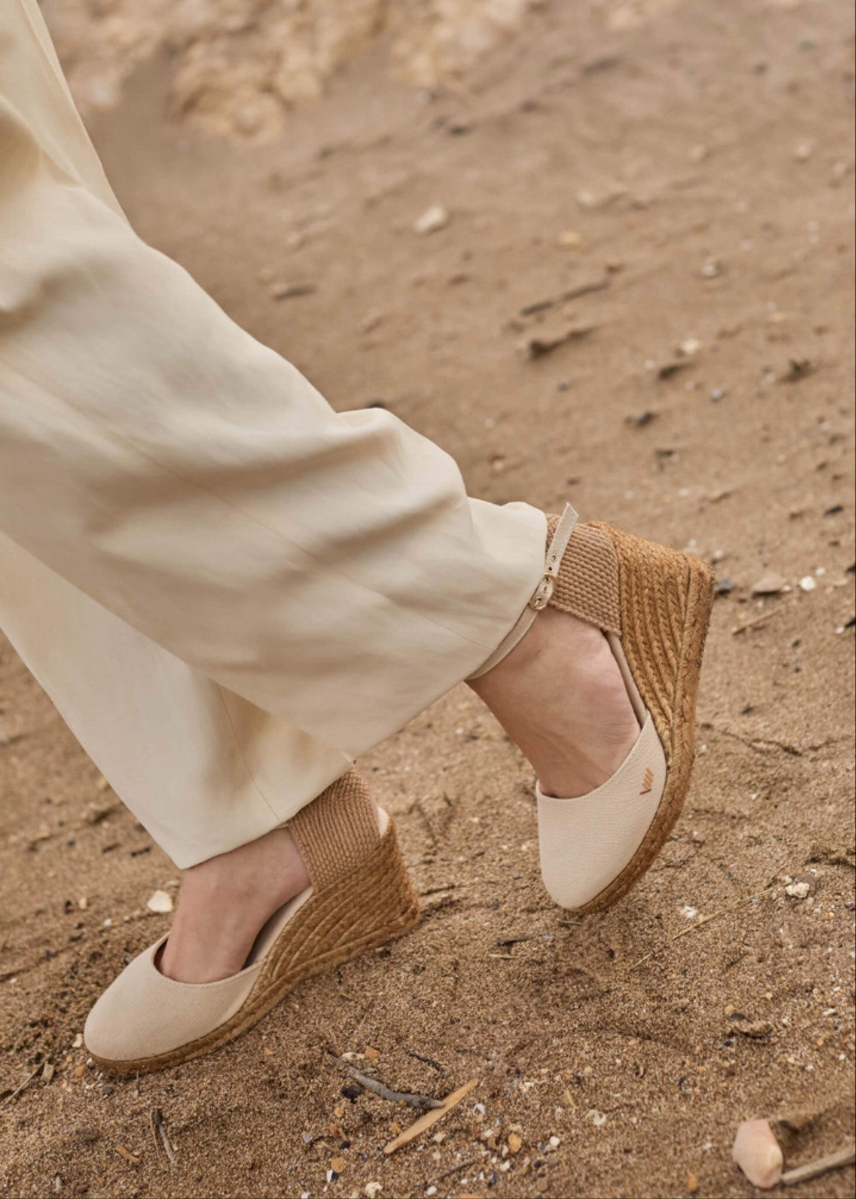 Beige espadrille wedges worn on model, walking on sandy ground in a natural setting.