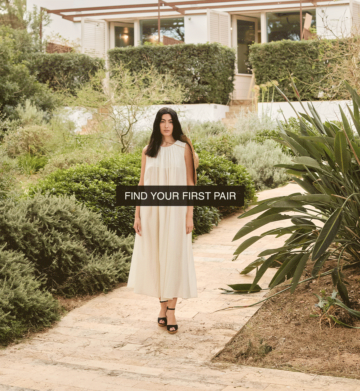 Woman in a flowing neutral dress walking through a Mediterranean garden wearing black espadrille sandals, highlighting effortless summer style in motion.