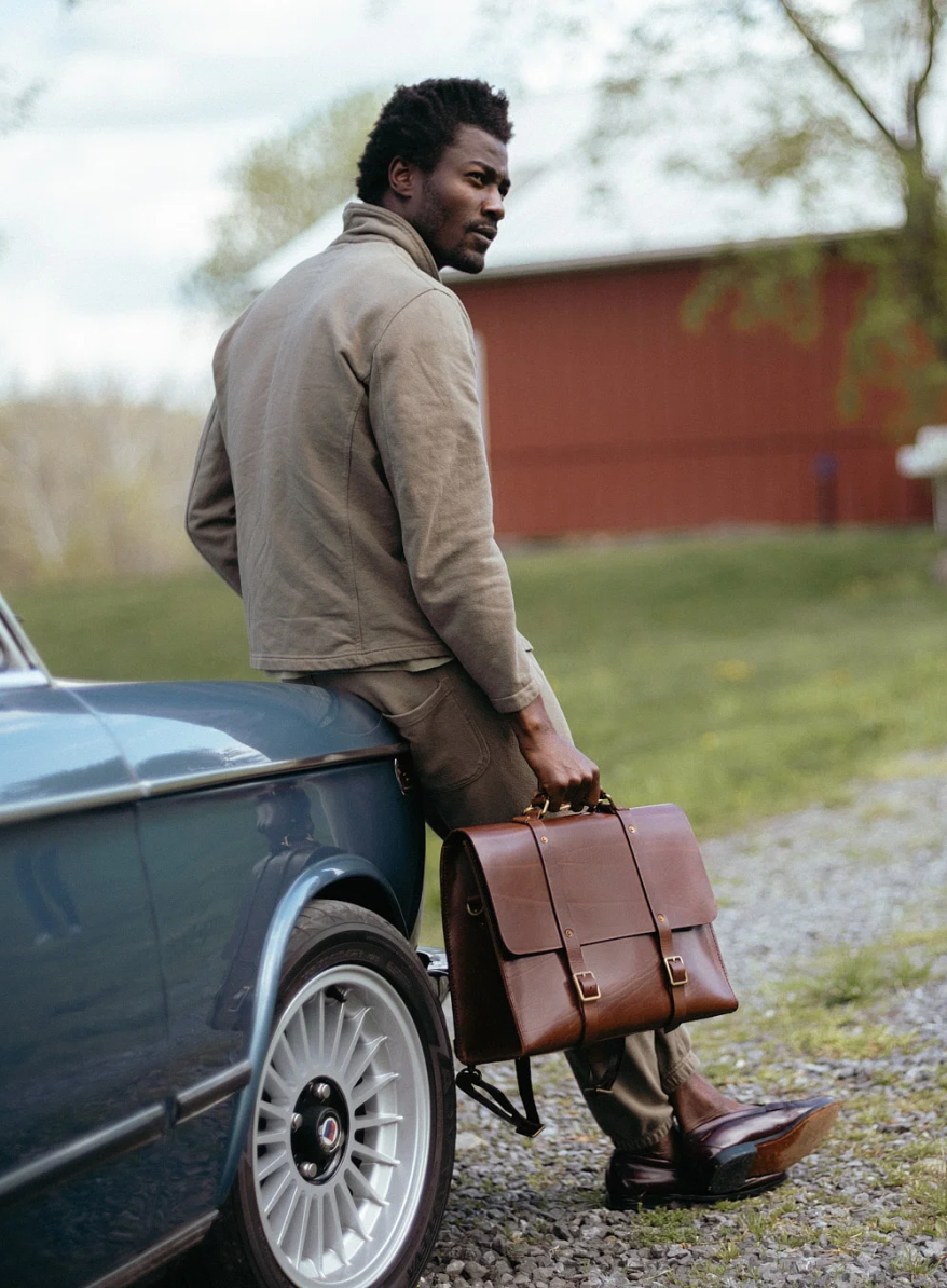 Man in a tan jacket leaning against a vintage blue car on a gravel road, holding a cognac leather satchel briefcase with buckle straps, a red barn visible in the background