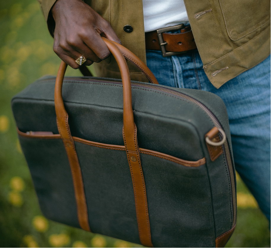 Person carrying a dark green waxed canvas briefcase with leather handles, casual outdoor setting with soft natural light