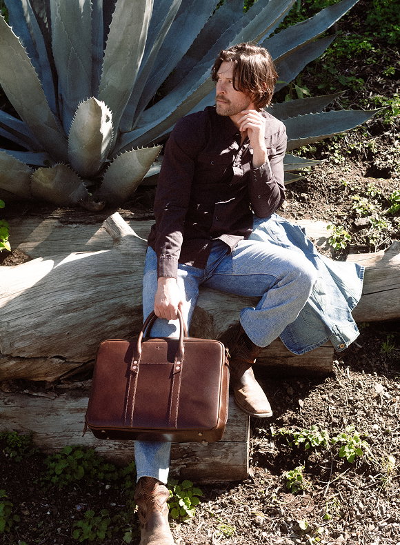 Man sitting on a fallen log next to a large agave plant, holding a structured brown leather briefcase, natural outdoor setting with soft sunlight