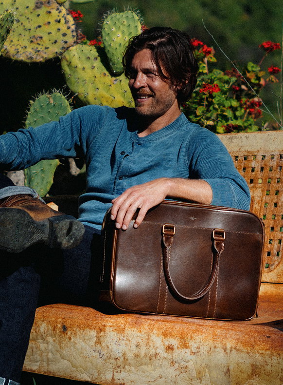 Man sitting outdoors on a rustic bench surrounded by cactus plants, holding a large brown leather briefcase on his lap, relaxed lifestyle moment in warm sunlight