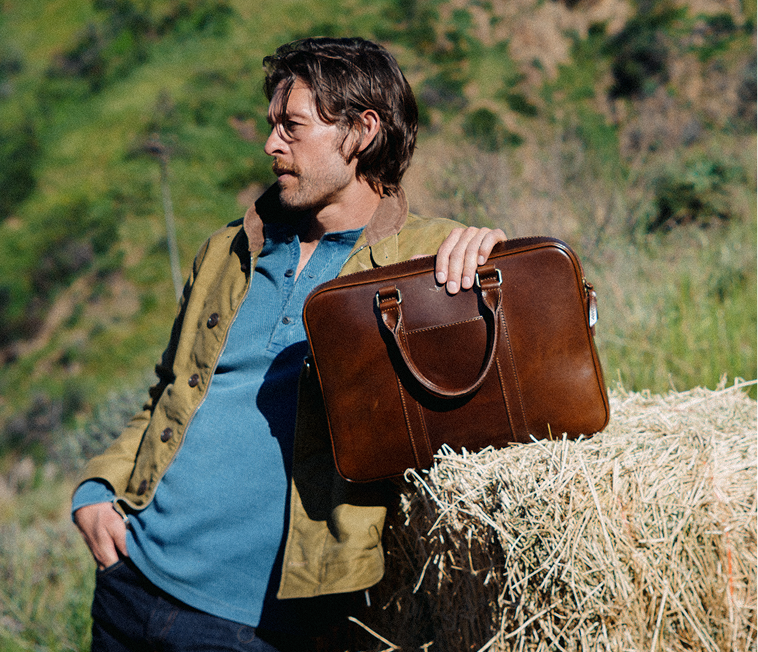 Man in a tan jacket and blue henley leaning against a hay bale outdoors, holding a structured brown leather briefcase by its top handles