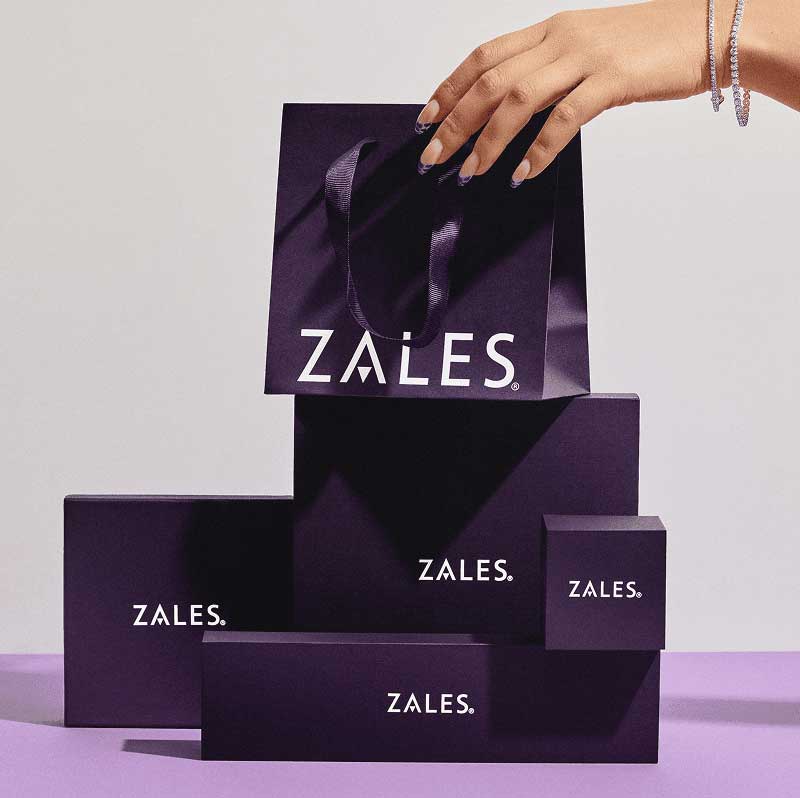 Model’s hand with layered in tennis bracelets, reaching for Zales purple shopping bag & various size boxes.