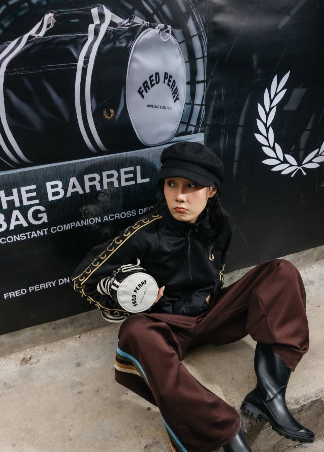 Female model sits on the side of the road on a walkway in front of a large Fred Perry barrel bag poster and wears the black and champagne track jacket while holding a mini barrel bag under her arm