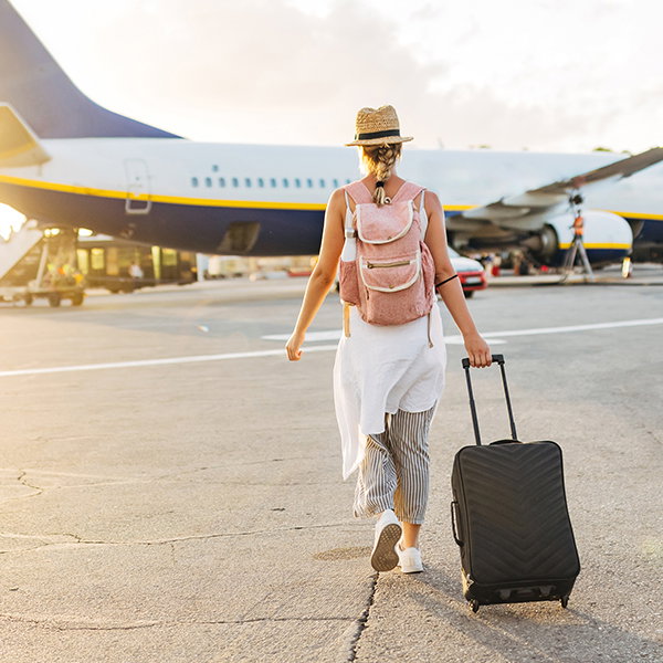Frau mit Strohhut, Rucksack und Rollkoffer geht auf ein Flugzeug zu