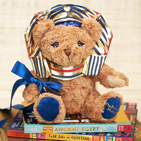 Teddy bear inspired by ancient Egyptian pharaohs with a blue ribbon, sitting on a stack of children’s books. - Shop Children Collection.