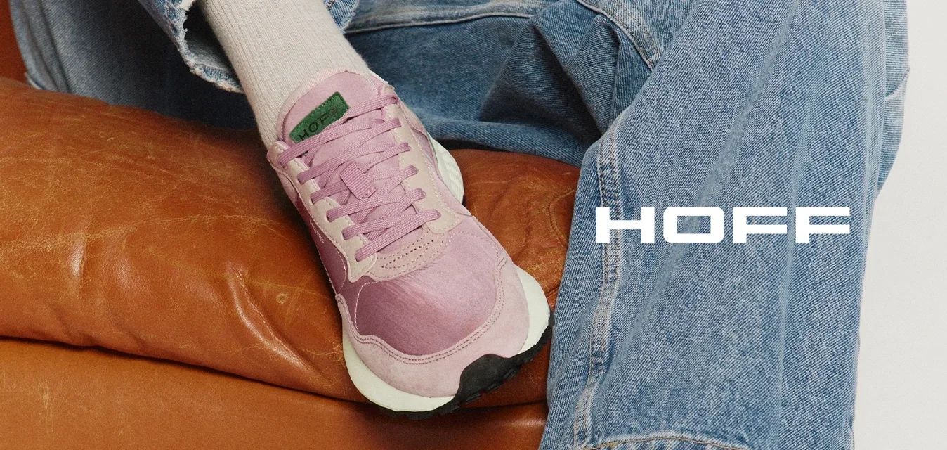 Persona con zapatillas deportivas color rosa claro, calcetines beige y jeans azules sentada en un sofá de cuero marrón con las piernas cruzadas.