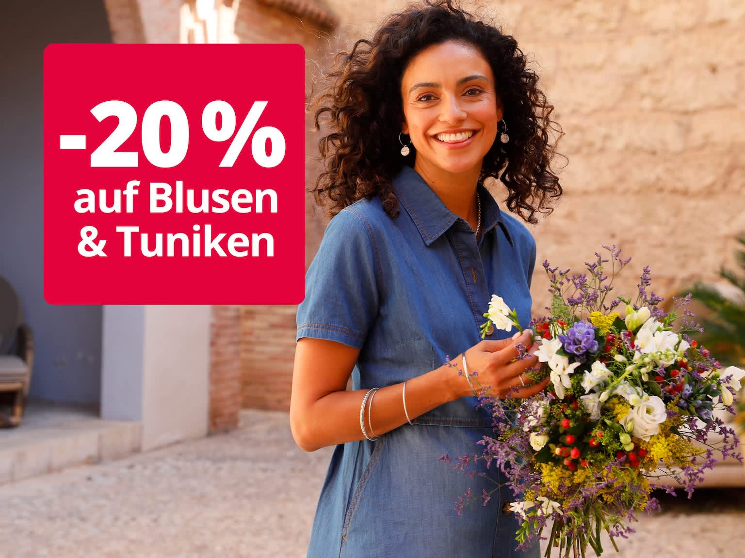 Das Bild zeigt eine Frau mit lockigen Haaren in einem blauen Kleid, die einen bunten Blumenstrauß vor einer mediterranen Steinmauer hält. Auf roter Fläche steht der Text '-20 % auf Blusen & Tuniken'.