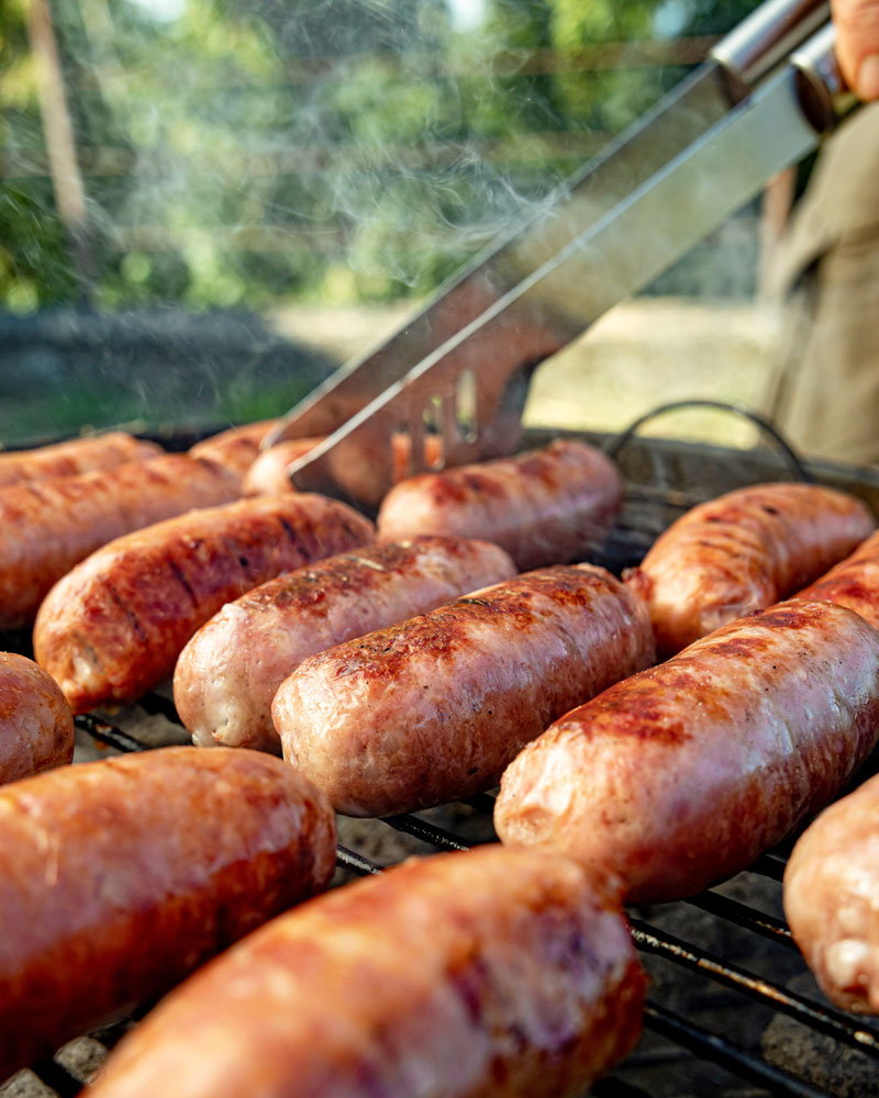 Gegrillte Salsiccia (Würstchen) auf einem Rost, die mit einer Grillzange gewendet werden, im Hintergrund aufsteigender Rauch.