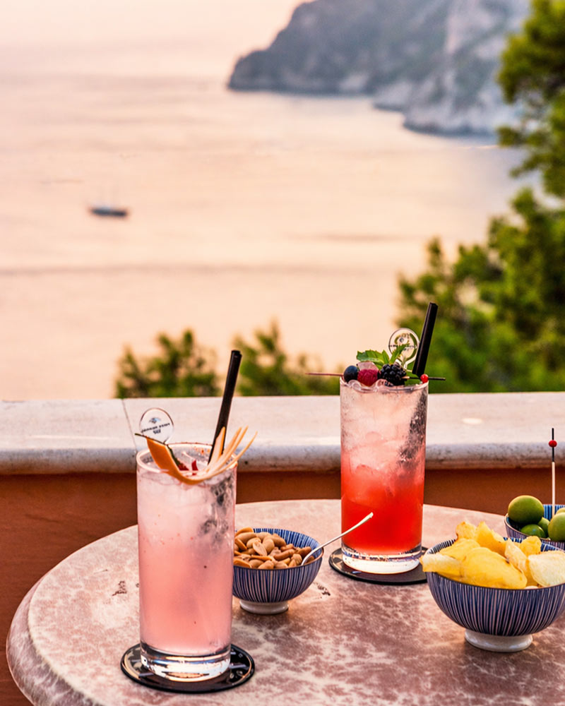 Zwei erfrischende Aperitif-Getränke auf einem Tisch mit Snacks wie Nüssen und Obst, serviert auf einer Terrasse mit Blick aufs Meer.
