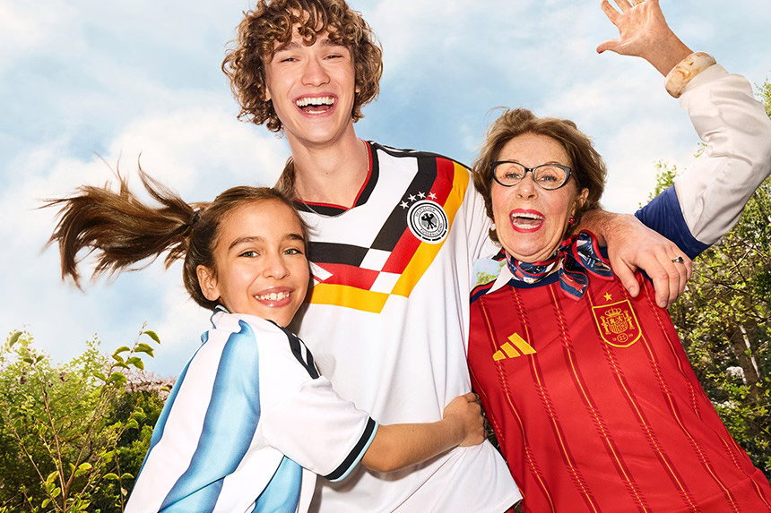 Drei Personen stehen draußen, zwei umarmen die mittlere Person. Sie tragen Fußballtrikots aus Deutschland, Argentinien und Spanien. Alle freuen sich und jubeln.