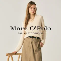 Femme portant un pull crème, une jupe midi beige, une ceinture noire et des mocassins noirs, debout à côté d'un support pliant en bois sur un fond uni.