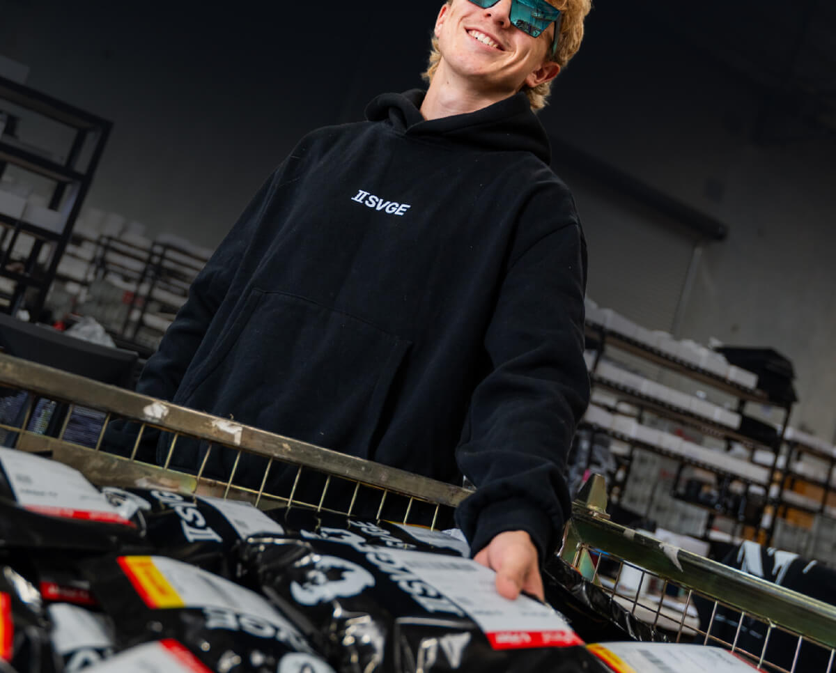 Young blonde person in black Svge hoodie and blue sunglasses sorting items in warehouse with metal cart