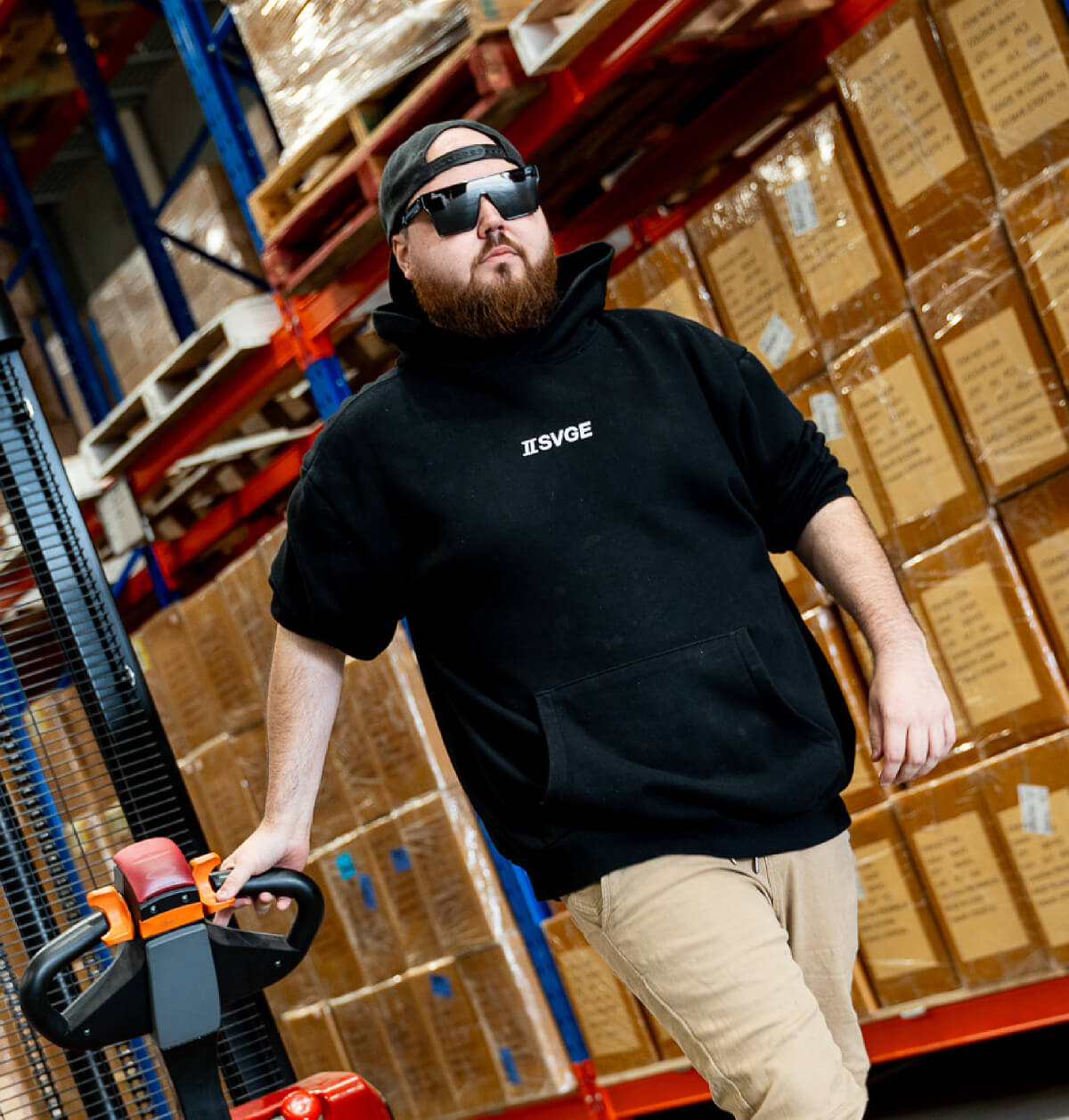 Warehouse worker in black Svge hoodie and dark sunglasses operating equipment among shelves of boxes
