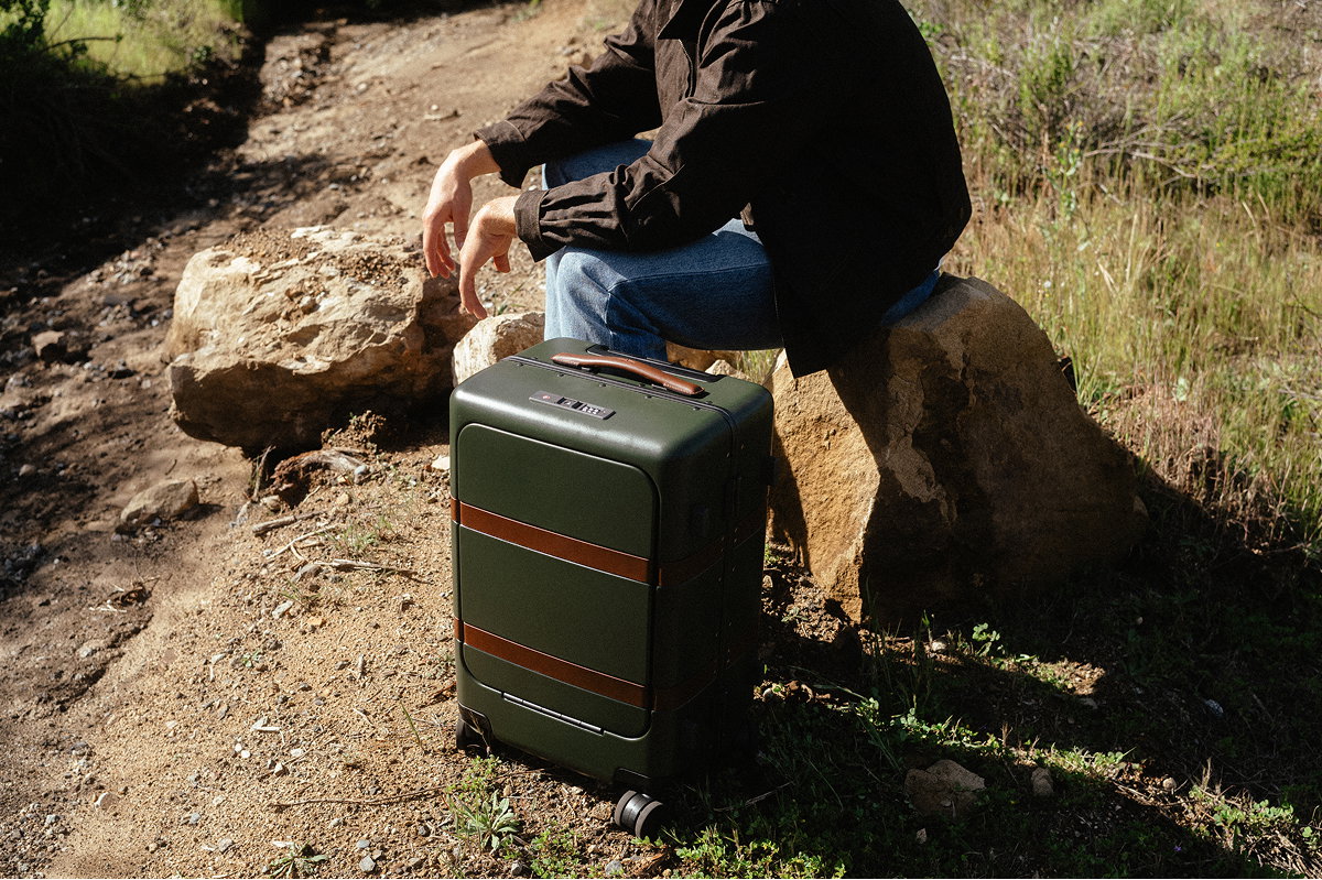 Traveler sitting on a rock beside a dark green hard-shell carry-on suitcase with leather accents on a sunlit dirt trail.