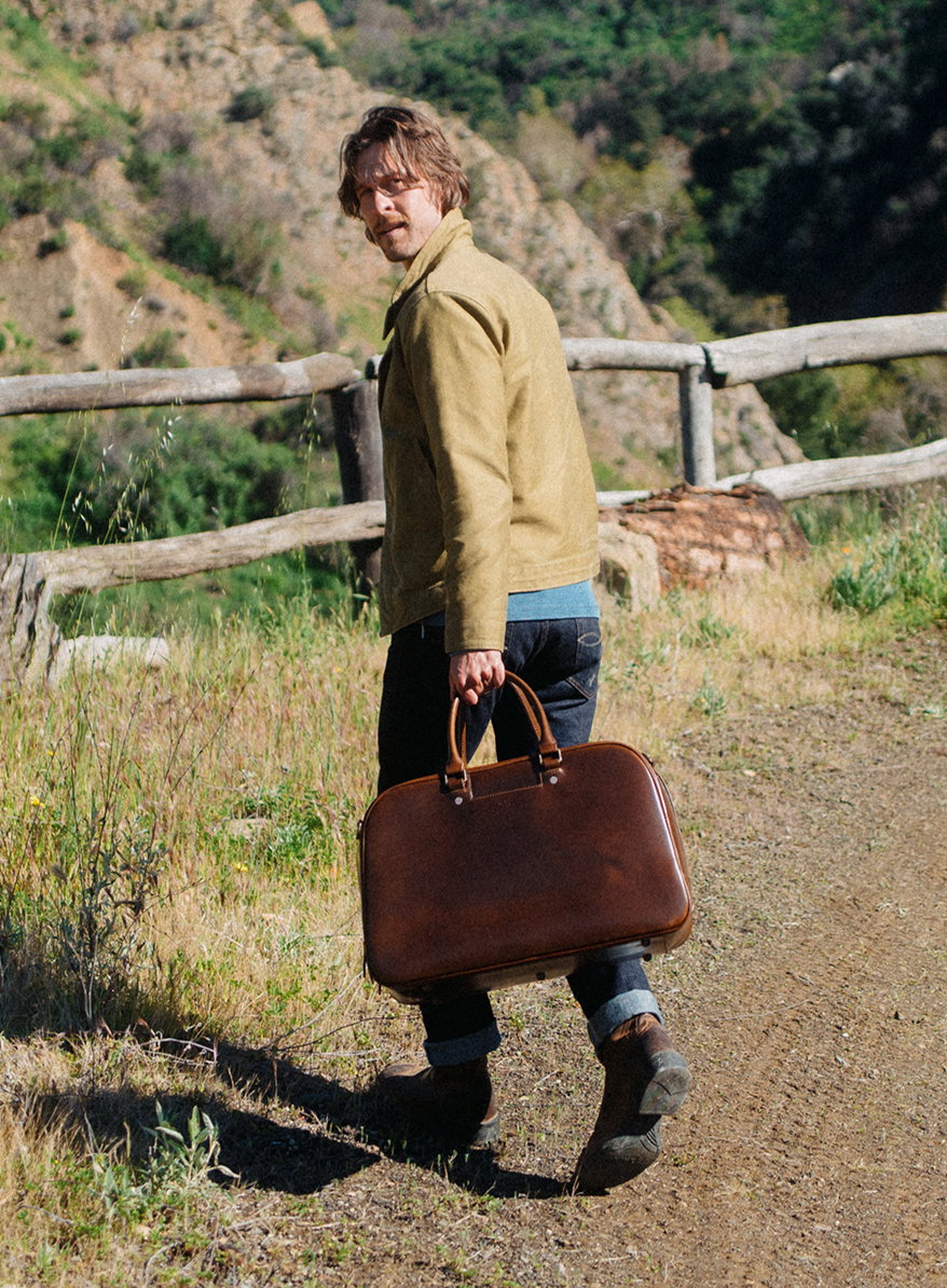 Man walking along a mountain trail carrying a brown leather briefcase, wearing a tan jacket and jeans in a rugged outdoor setting.