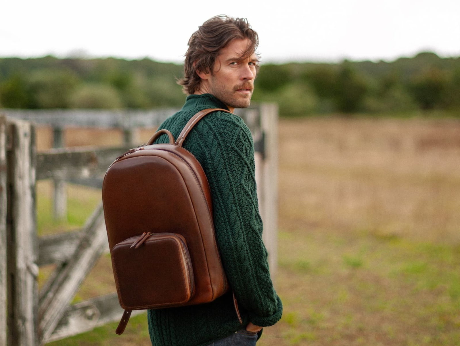 Man in a light blue shirt leaning into a horse stall to greet a horse, wearing a sleek brown leather backpack with a front zip pocket