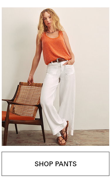 A woman wearing a sleeveless orange top and wide-leg white pants stands next to a woven chair, with one hand in her pocket. She is also wearing sandals and a brown belt. Text below reads "SHOP PANTS.